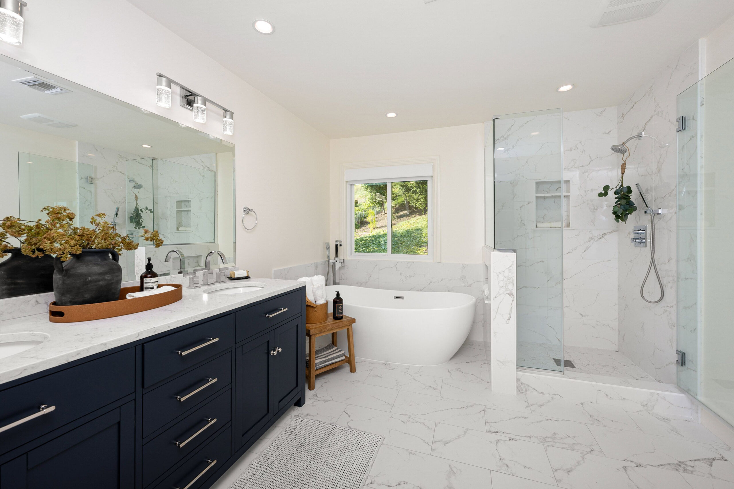 A modern bathroom interior with a freestanding tub, glass shower, double vanity with dark blue cabinets, and a large mirror.