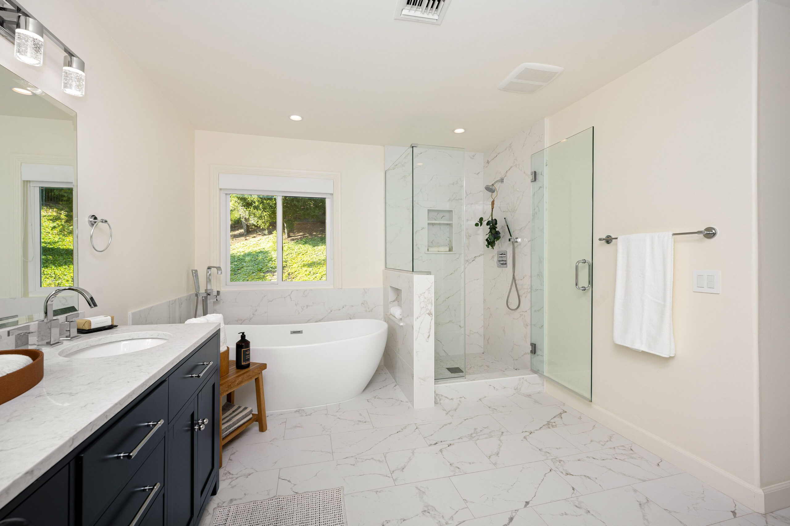 A modern bathroom with marble flooring, a freestanding bathtub, and a glass-enclosed shower with a window view.