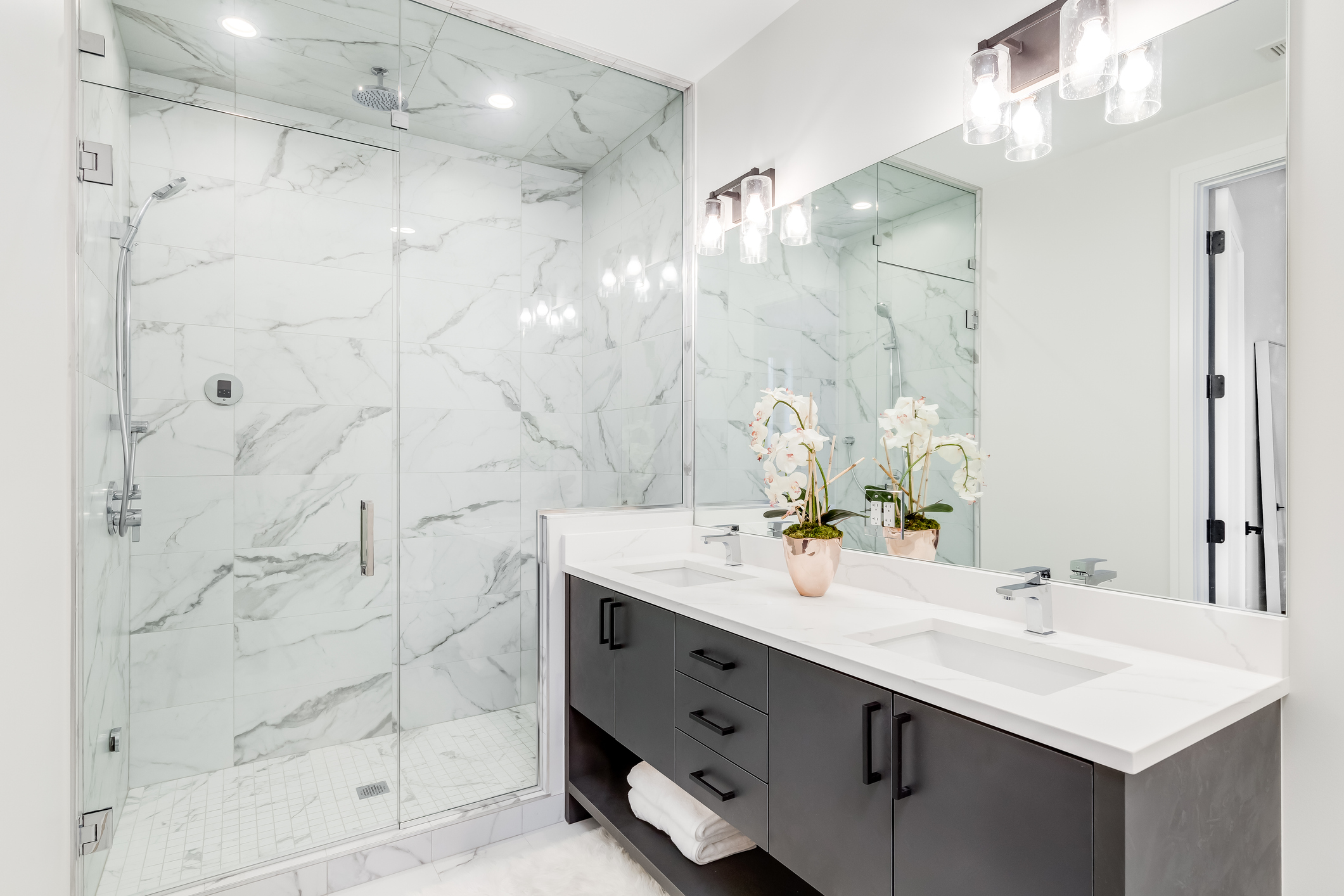 A beautiful bathroom with a dark vanity and white granite counter top. A plant and towels sit on the vanity. The shower is lined with marble tiles. Lights on.