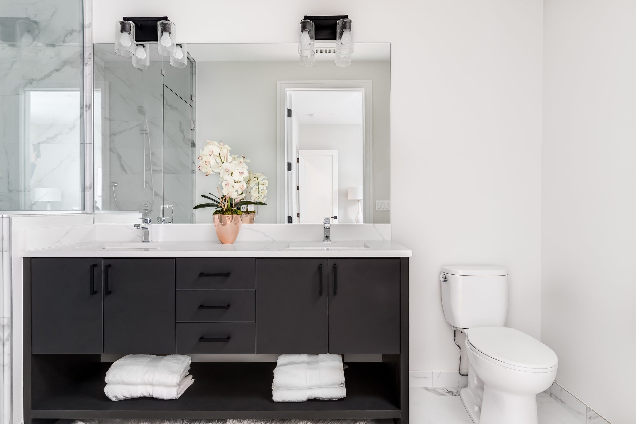 A beautiful, modern bathroom with a dark vanity and white granite counter top. A small plant and towels sit on the vanity. The shower is lined with marble tiles.