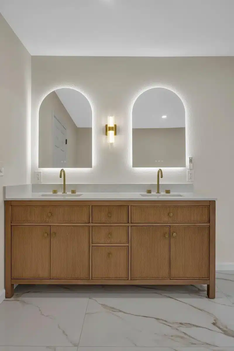 Modern bathroom with wooden dual vanity, arched LED-lit mirrors, brass fixtures, and marble flooring