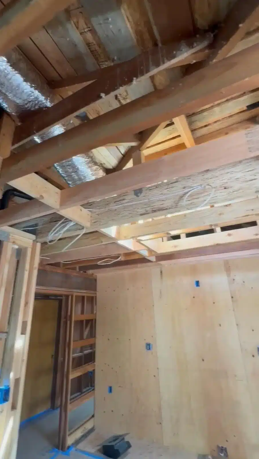 Exposed wood ceiling beams, foil-wrapped ducts, loose wiring, and plywood wall sheathing in an unfinished interior.