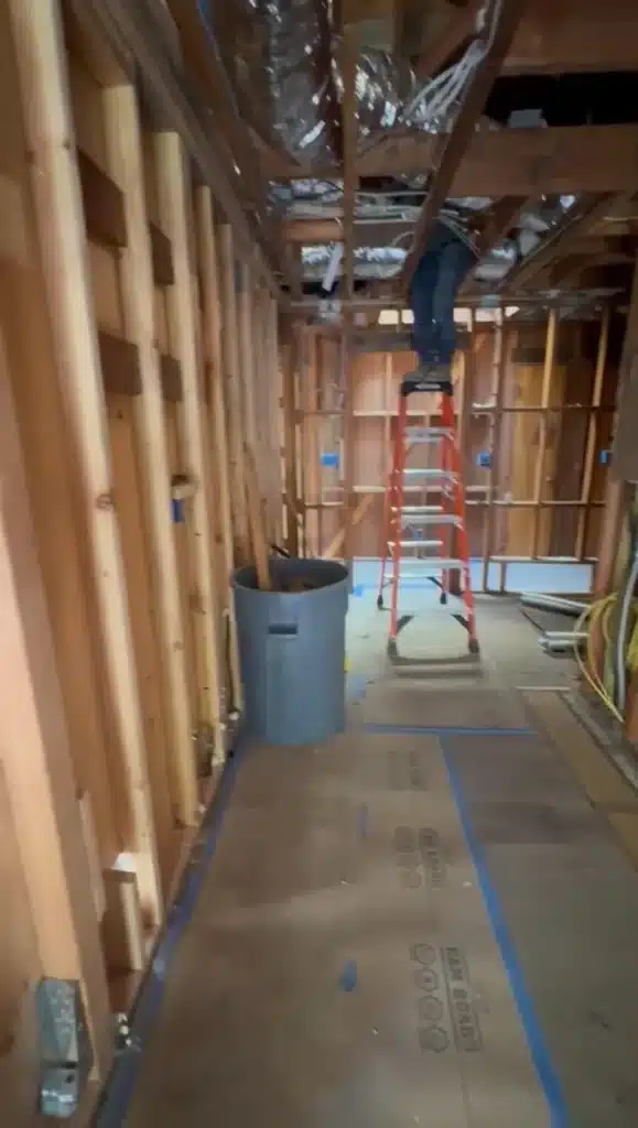 Wood-framed hallway with exposed ceiling ducts, a red ladder, gray debris bin, and taped floor protection.