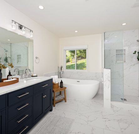 A modern bathroom interior with a freestanding tub, glass shower, double vanity with dark blue cabinets, and a large mirror.