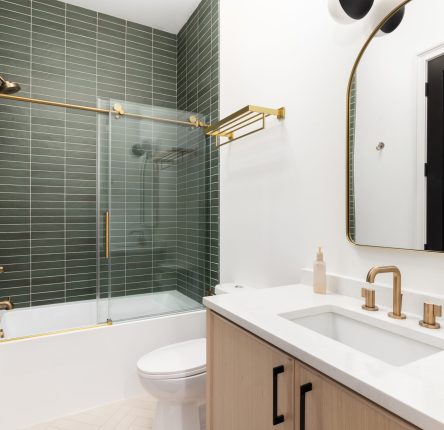 A bathroom with a wood vanity cabinet, gold hardware and faucet, and a shower with green subway tiles.